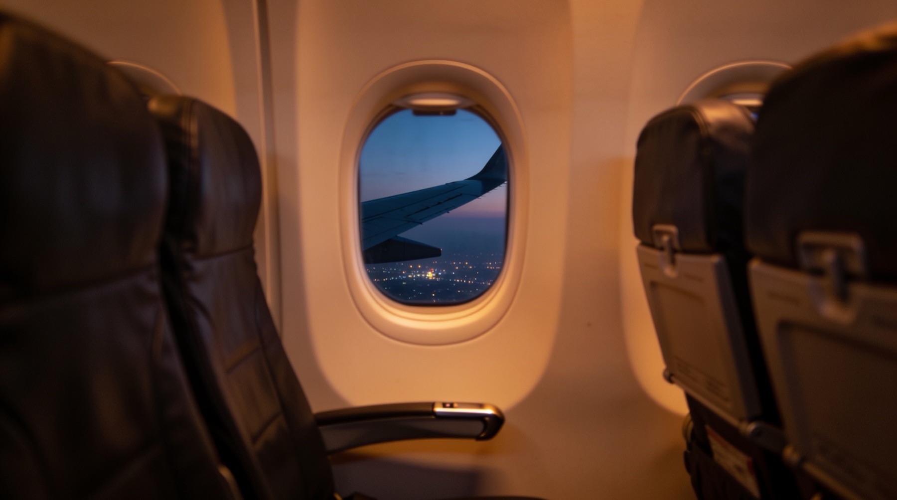 The view from an airliner window seat at dusk, a few rows forward of the wing — amber cabin light, the wing stretching into blue-grey twilight over a faint carpet of city lights
